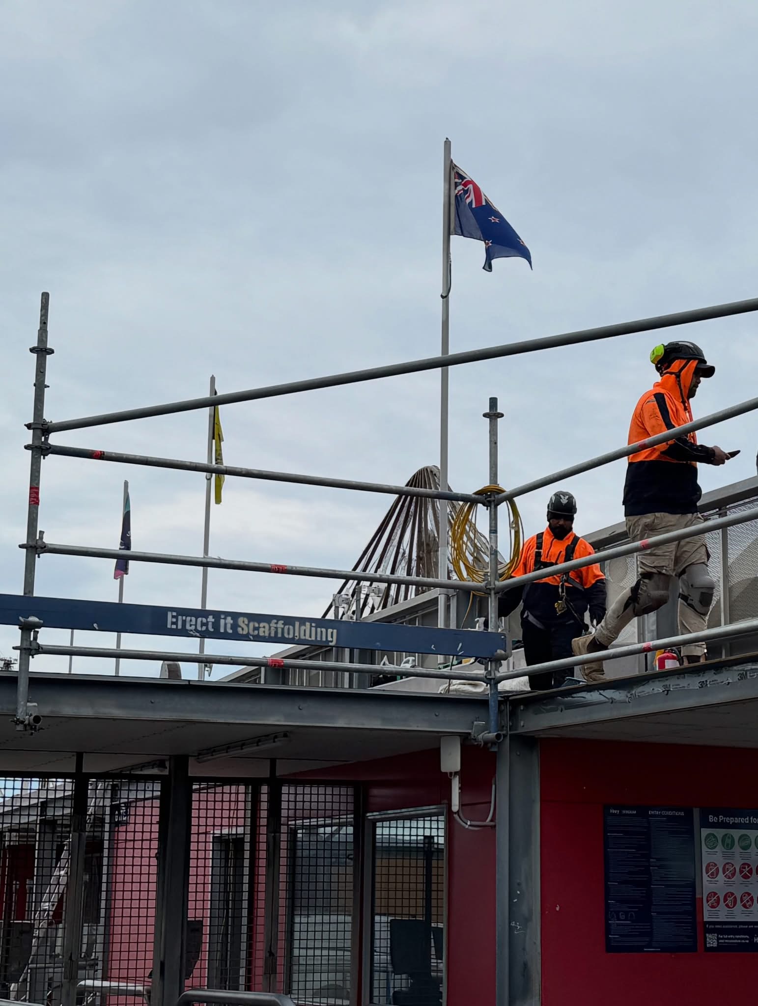 Erect It Scaffolding crew working at height on edge protection at HNRY Stadium, Wellington — New Zealand flag flying above the completed roofline installation