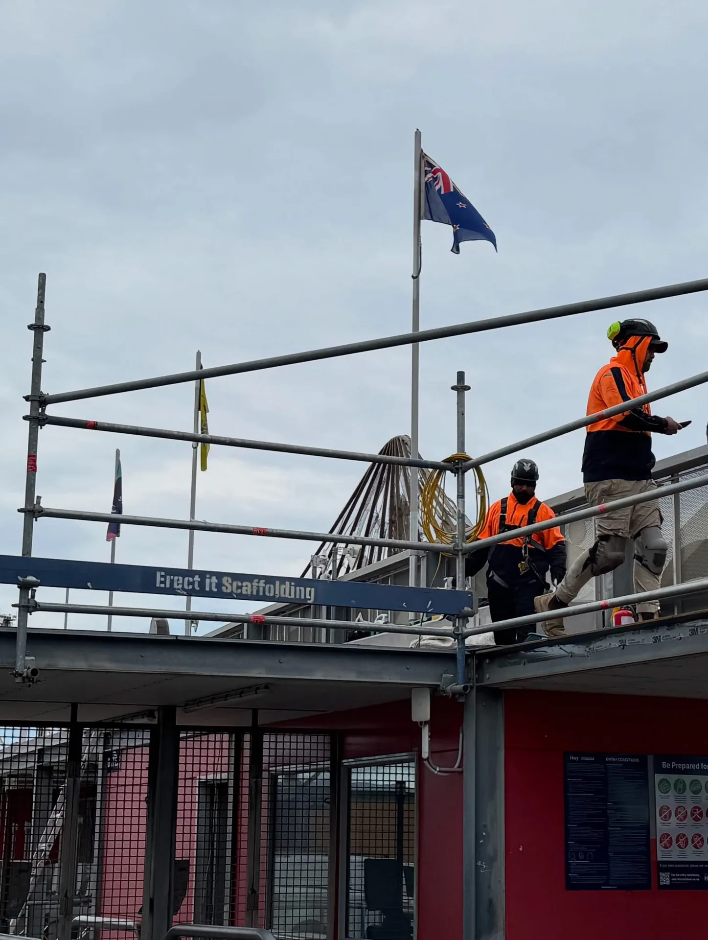 Erect It Scaffolding crew working at height on edge protection at HNRY Stadium, Wellington — Erect It Scaffolding branded board and New Zealand flag visible above the rooftop installation