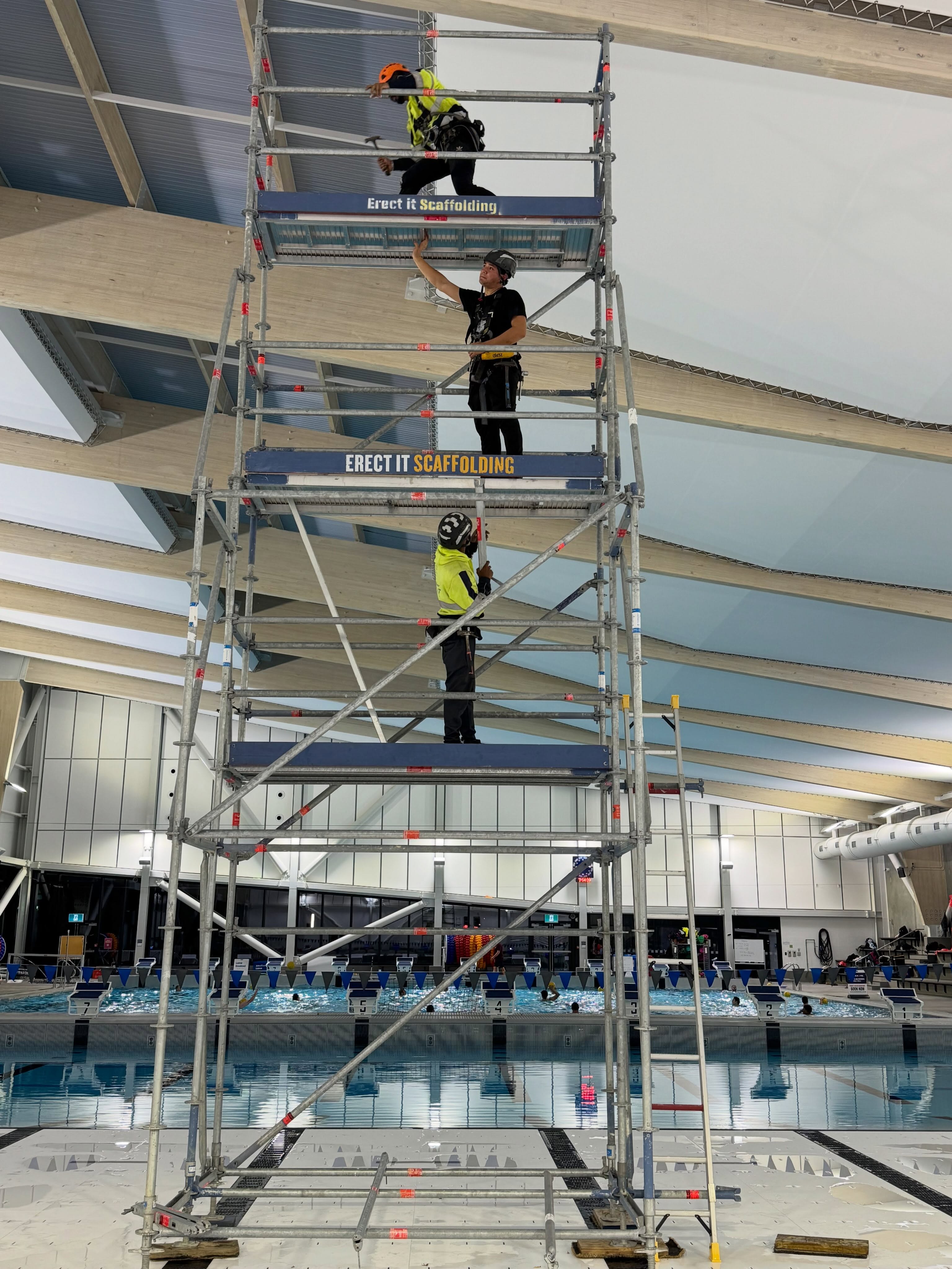 Erect It Scaffolding crew working at height on a custom scaffold tower inside the Naenae Pool aquatic centre in Lower Hutt — installing a goal system at ceiling height above the competition pool