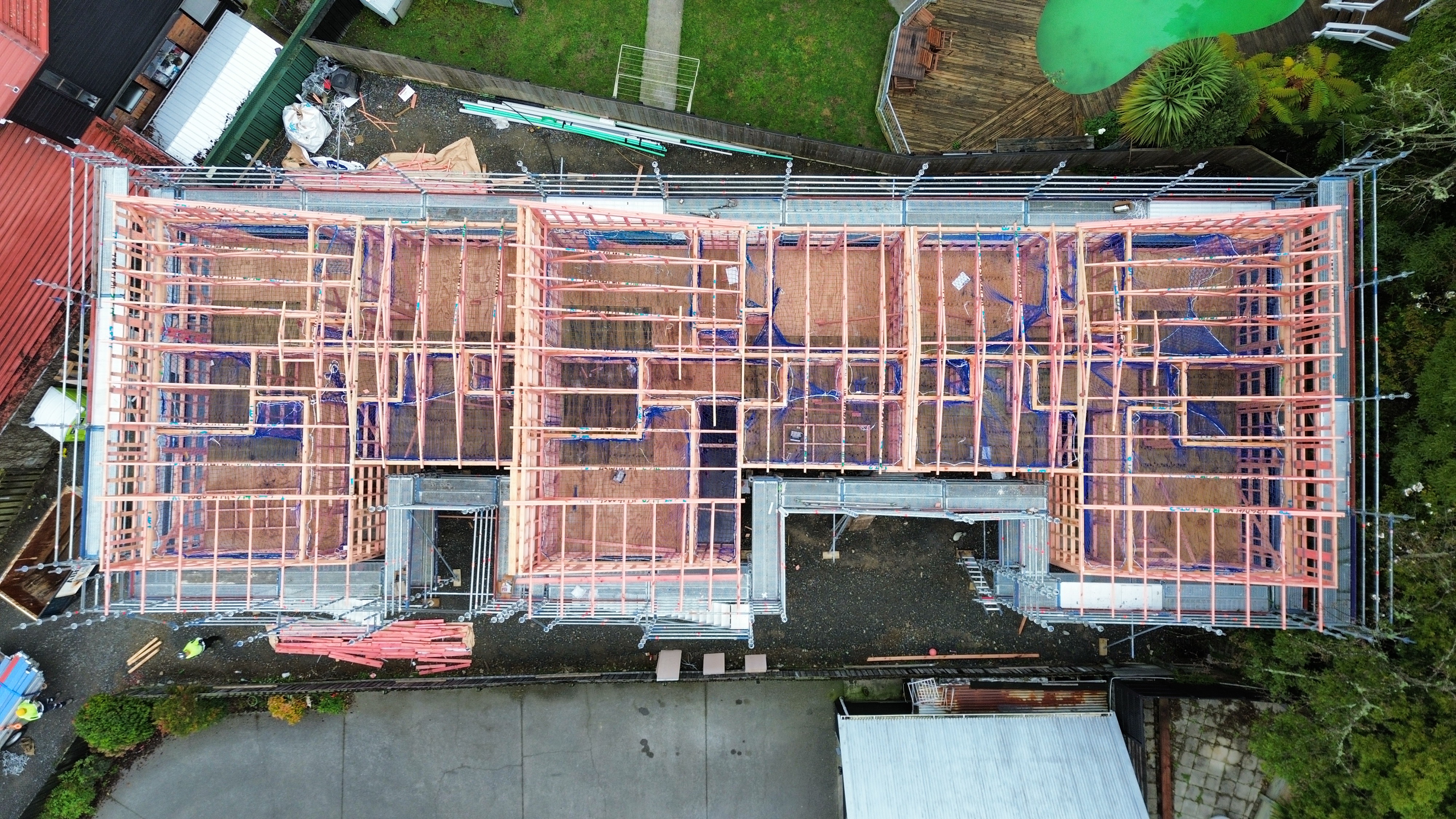 Aerial drone top-down view of new residential build at framing stage in Trentham, Upper Hutt — full scaffold perimeter visible wrapping the timber frame with safety nets and scaffolding bracing clearly visible